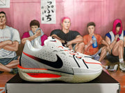 Side profile of a white Nike basketball sneaker with a black Swoosh, orange-red trim, perforated panels, and speckled midsole.