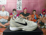 Side view of an off-white Nike basketball sneaker with gray accents, raised geometric textures, and a black Swoosh.