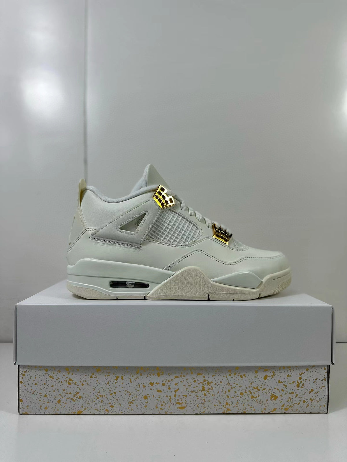 Side profile of white Jordan 4 sneakers with gold-tone lace hardware displayed on a shoebox.
