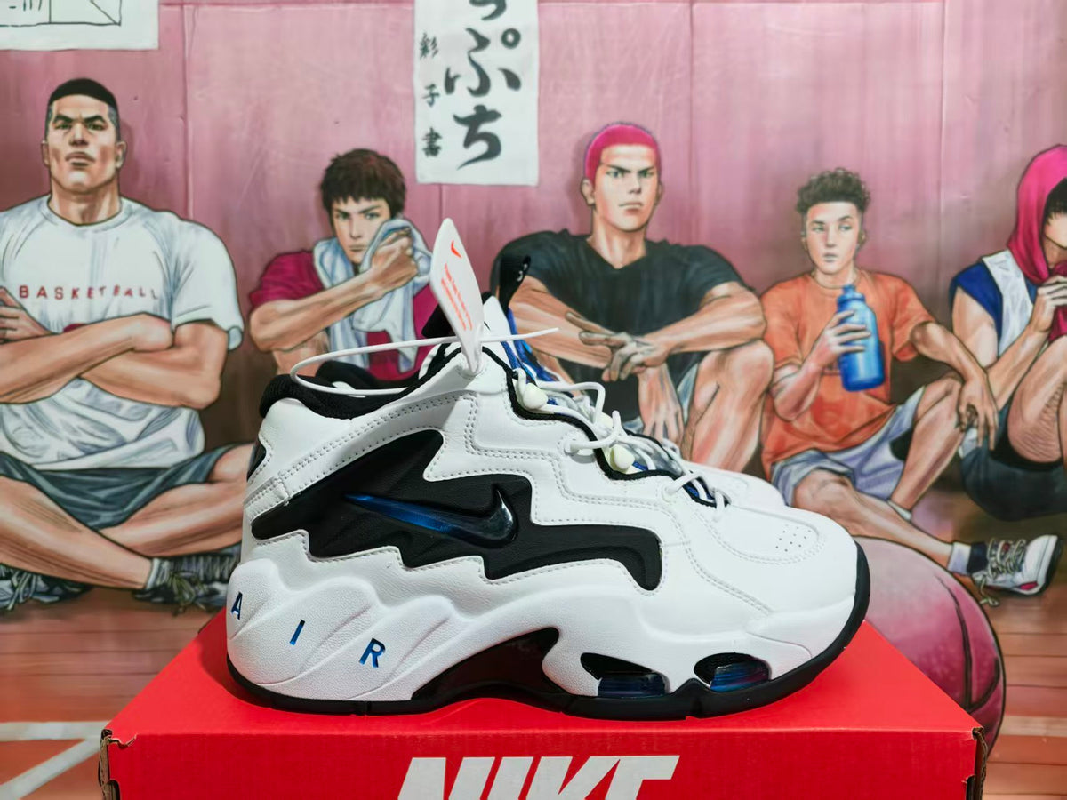 Side view of white Nike basketball-style sneakers with black and royal blue accents displayed on a red Nike box.