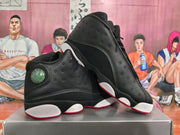 Angled view of two black high-top basketball sneakers highlighting perforated side panels, layered construction, white outsole sections, and red accents.