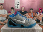 Side profile of a gray Nike basketball sneaker with a white Swoosh, blue speckled support panel, and blue outsole.