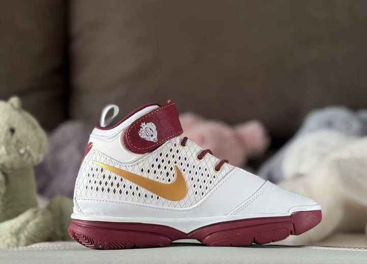 Side view of a white high-top kids' sneaker with a metallic gold Swoosh, deep red ankle strap, and deep red outsole.