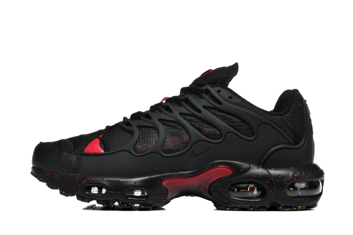 Side view of black Nike Air Max Plus-style sneakers with red accents, a wavy side cage, and cushioning windows in the sole