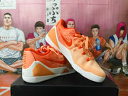 Pair of orange and white Nike basketball sneakers displayed on a black box, showing the side Swoosh, perforated panels, and Kobe-style heel details.