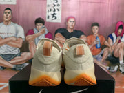 Rear view of cream Nike sneakers with orange pull tabs and translucent orange outsoles.