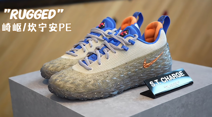 Pair of gray Nike sneakers with blue accents, orange lining, textured uppers, gray-and-white laces, and dark patterned outsoles displayed on a platform; the image includes “RUGGED” and “ST. CHARGE” text.