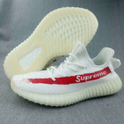 White low-top sneaker with a red side stripe, visible “Supreme” lettering, patterned upper, and translucent ribbed sole.