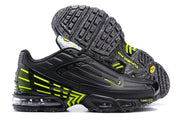 Side view of a black and volt Nike Air Max Plus sneaker beside its speckled outsole.