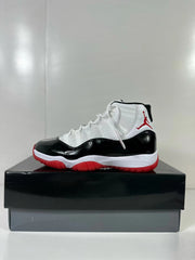 Side view of black, white, and red Jordan Air Jordan 11 Retro sneakers displayed on a black shoebox.