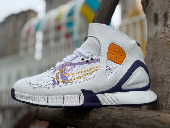 Side view of a white high-top Nike basketball shoe with purple map graphics, gold Swoosh, and orange side panel