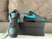 Front three-quarter view of the sneakers beside a black shoe box, showing black laces, perforated toe panels, and turquoise tongue and side logos.