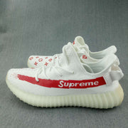 Side view of multiple white sneakers showing the red stripe, printed motifs, rear pull tabs, and ribbed soles.