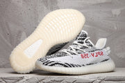 Side view of black-and-white Yeezy 350 V2 sneakers with a light ribbed sole; one shoe is lifted to show the outsole.