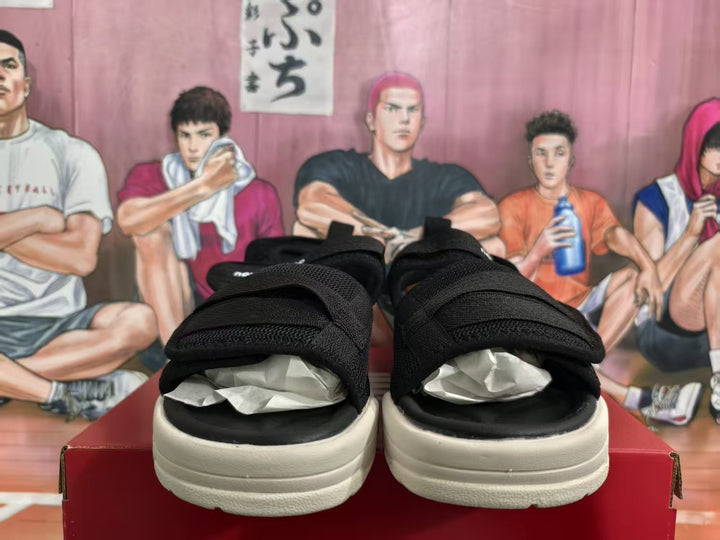 Pair of black New Balance sandals viewed from the front, showing broad adjustable straps and off-white soles on a red box.