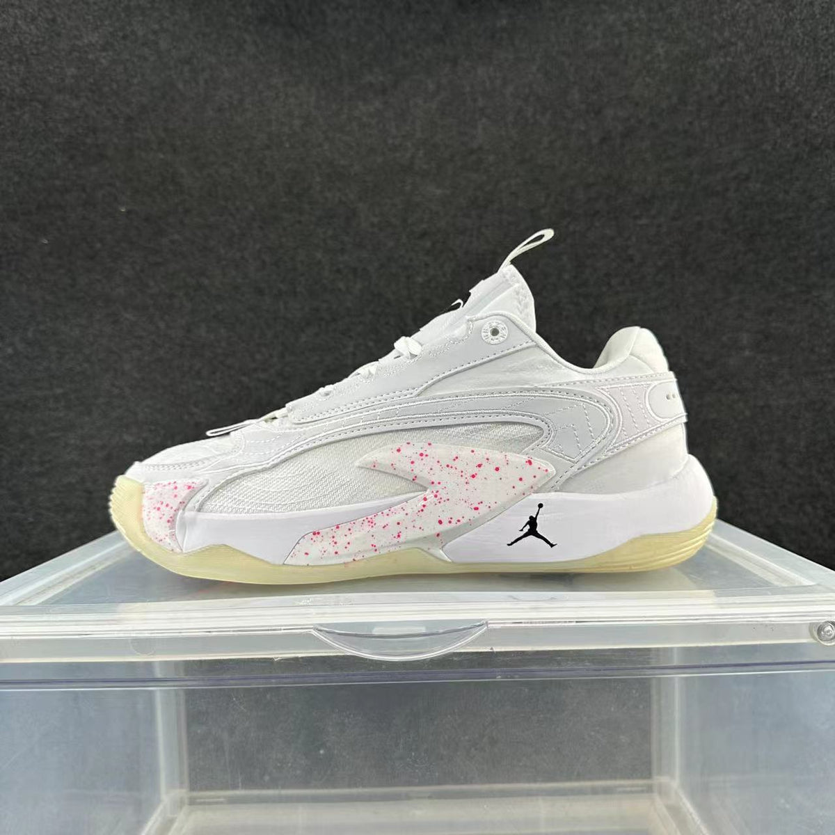 Side view of a white low-top sneaker with pink speckled accents, a black Jumpman-style logo, and a pale yellow outsole.