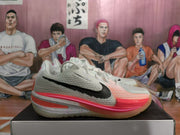 Side profile of a white Nike basketball sneaker with a black Swoosh, textured upper, coral and pink midsole accents, and translucent outsole.