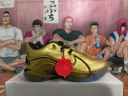 Side view of a metallic gold Nike basketball shoe with black trim and a red charm