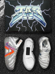 Grey and white Nike basketball sneakers with orange and pink side accents, displayed beside a black lightning-patterned box.