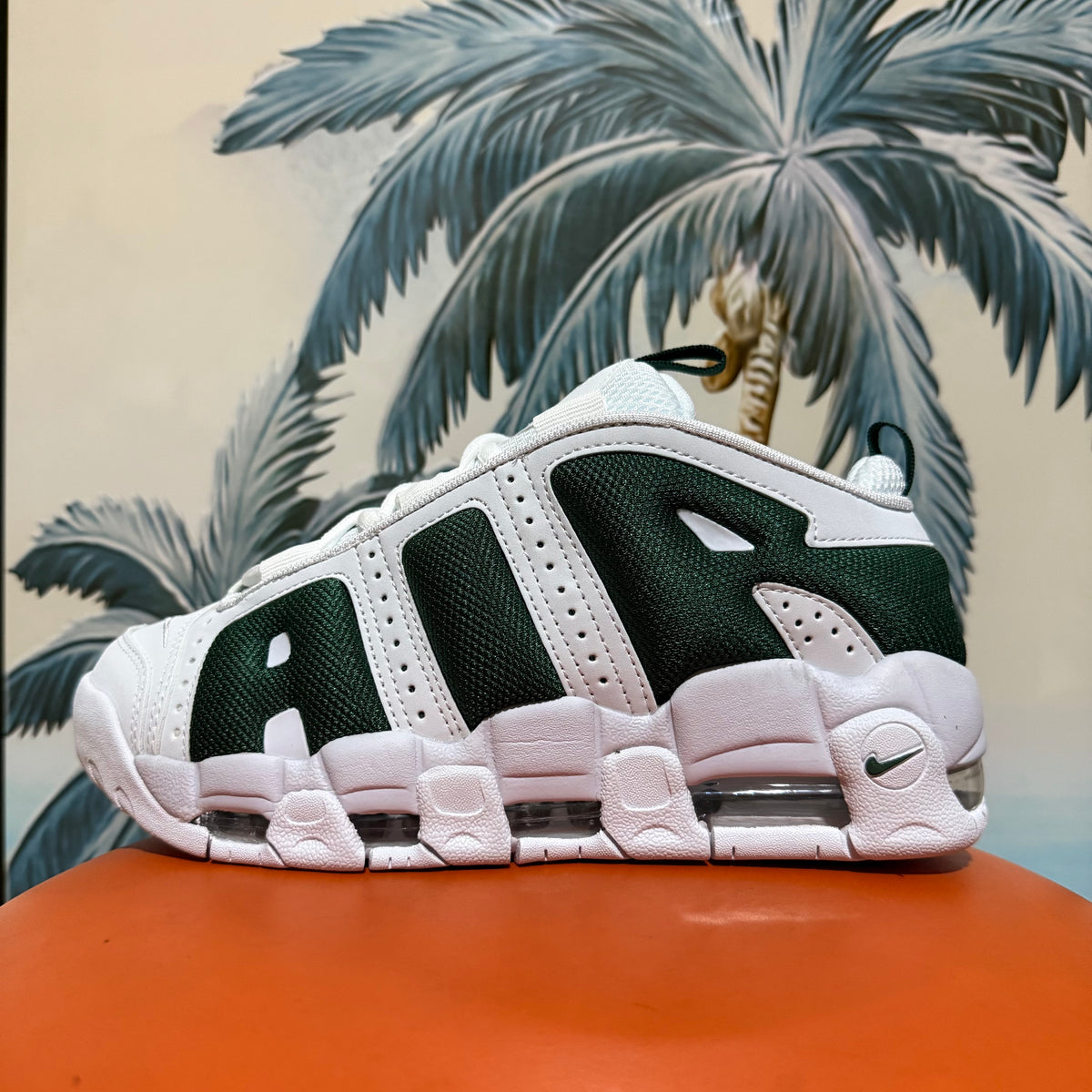 Side view of a white and dark green Nike sneaker displayed against a palm-leaf background.
