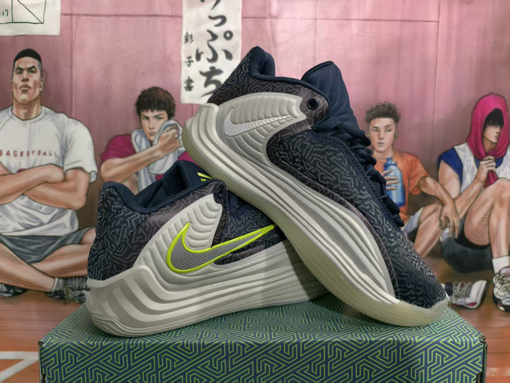 Pair of deep navy-gray Nike basketball sneakers displayed on a patterned box, showing the side Swoosh and layered light-colored soles.