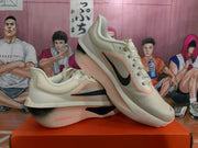 Angled view of two beige and black Nike running shoes showing the side swooshes, lace-up construction, pink accents, and ZoomX branding.