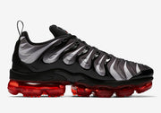 Side profile of a black and gray patterned Nike Air VaporMax Plus sneaker with a translucent red sole.