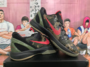 Angled view of paired camouflage Nike basketball shoes displayed on a box, showing red side branding and olive green heel panels.