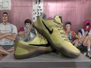 Angled view of gold-toned Nike basketball shoes displaying the black Swoosh, Kobe tongue emblem and layered upper.