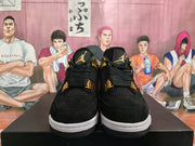 Front view of black sneakers with gold-toned tongue branding, lace details, and contrasting white midsoles.