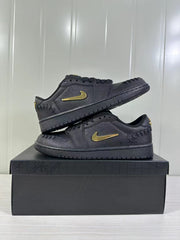Two black low-top sneakers stacked on a shoebox, showing the gold-tone side branding and low-profile shape.