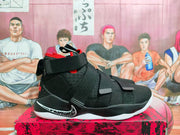 Side view of black Nike LeBron basketball shoe with red accents, white outsole, and wide straps.