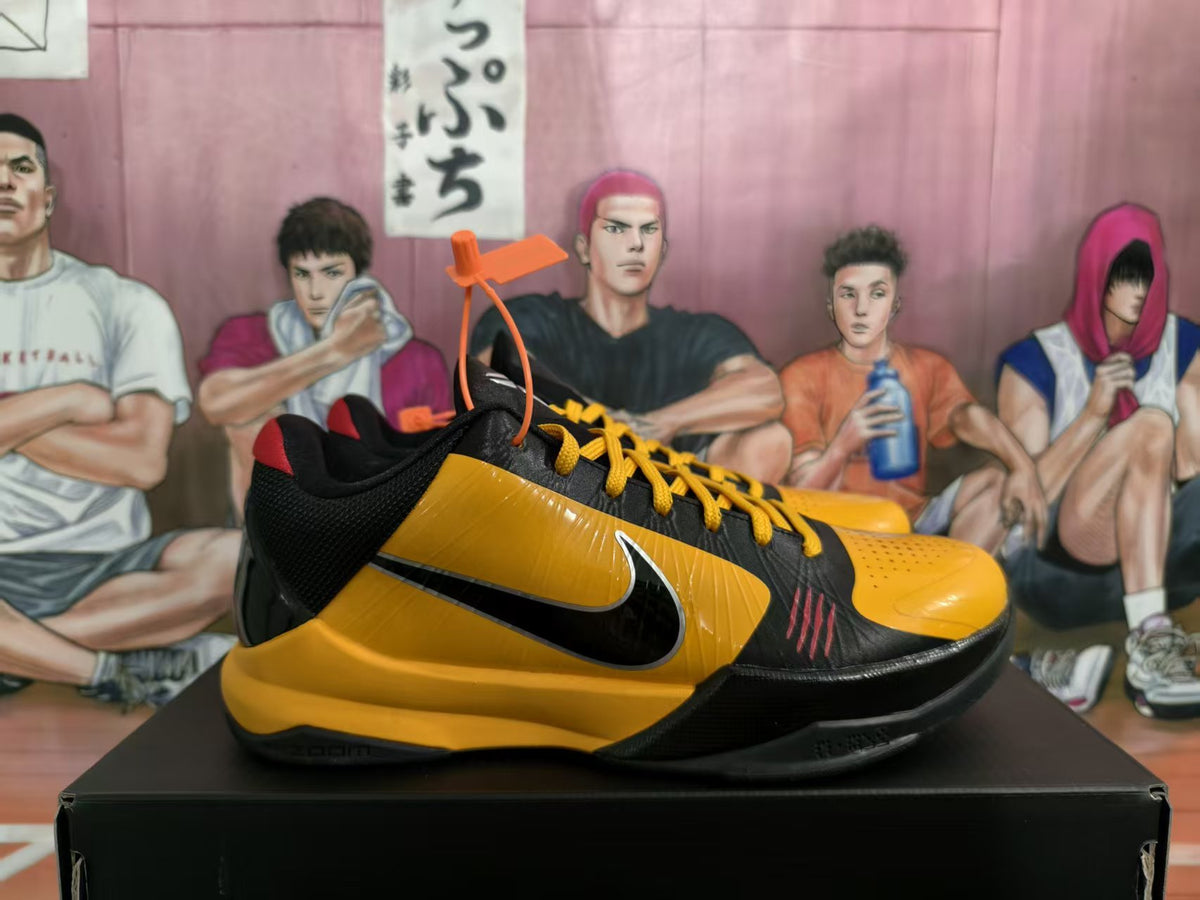 Side view of black and yellow Nike basketball-style sneakers with yellow laces and a visible Swoosh, displayed on a black box.