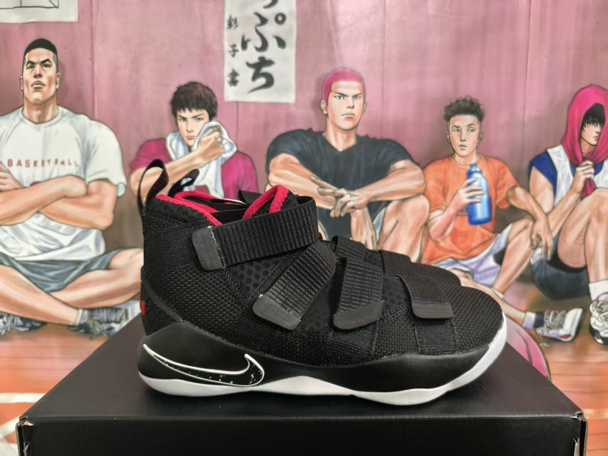 Side view of black Nike LeBron basketball sneakers with red lining, multiple straps, and a white outsole on a black shoebox.