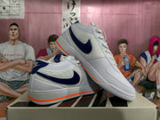 Two white Nike low-top sneakers shown from angled side views, featuring dark purple or navy branding details and orange outsole accents.