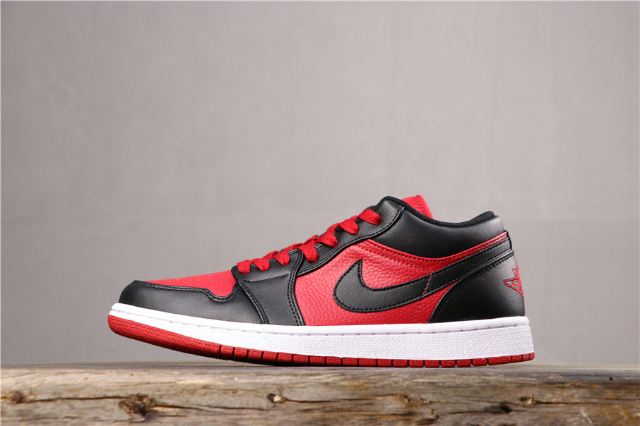 Side view of a black and red Jordan low-top sneaker with a white midsole and red outsole on a wooden surface.