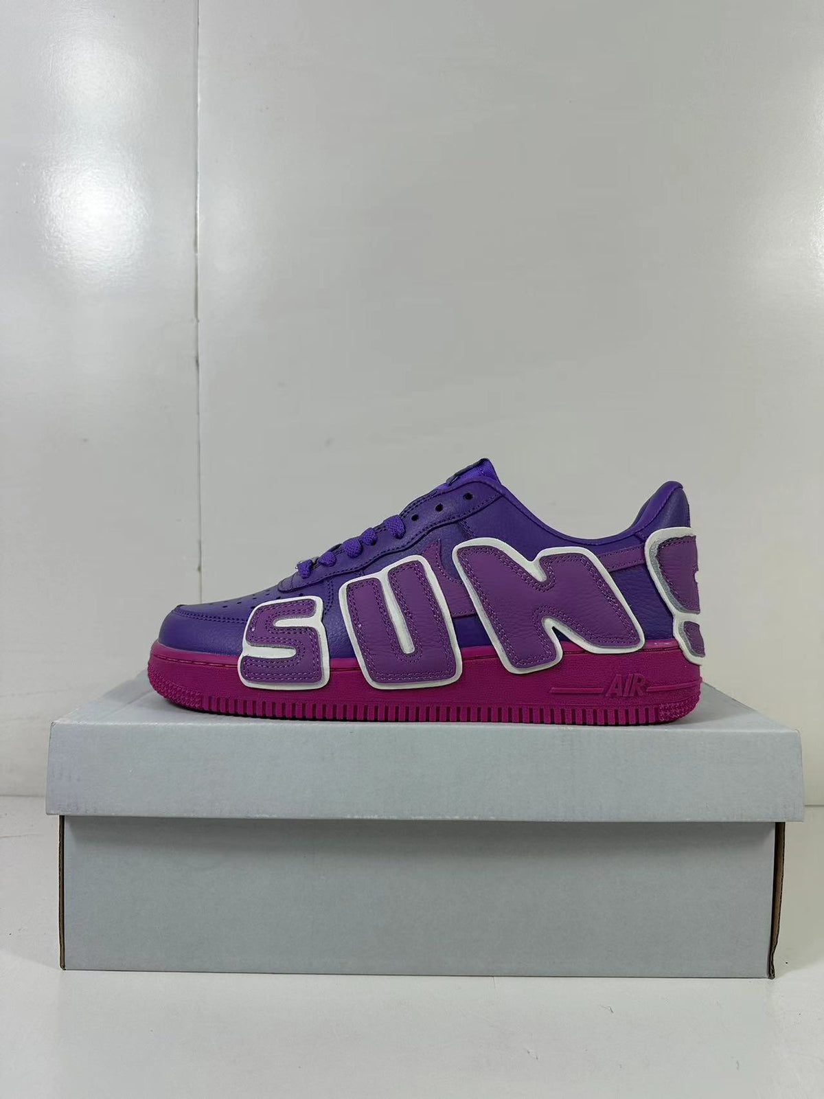 Side view of purple low-top sneakers with oversized AIR lettering and a magenta outsole on a box.