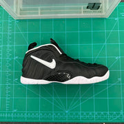 Side view of a black Nike Foamposite-style high-top sneaker with a white Swoosh and white outsole on a green measuring mat.