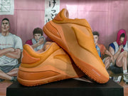 Pair of tangerine-orange slip-on sneakers displayed on a black box, showing the layered heel and textured outsole.