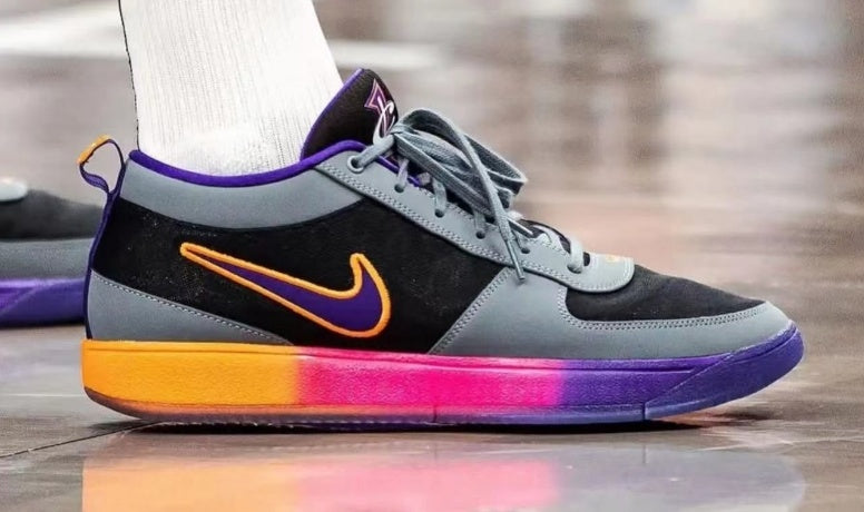 Side view of a gray and black Nike low-top sneaker with purple trim, an orange Swoosh, and an orange-to-pink-to-purple sole.