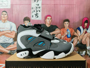 Side view of a gray and blue Nike basketball-style sneaker with an adjustable strap, layered panels, blue openings, and AIR lettering near the heel.