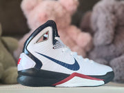 Side view of a white kids’ basketball shoe with a navy Swoosh, black heel and red midsole accent