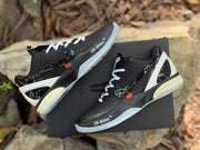 Pair of black basketball sneakers displayed on a black box, showing white laces, white heel structures, and contrasting side details.