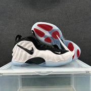 Pair of white and black sneakers displayed with the outsole facing upward, showing red traction sections and translucent rubber.