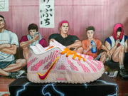 Side view of white Nike sneakers with pink textured detailing, orange laces, and a pink-and-yellow Swoosh on a black box.