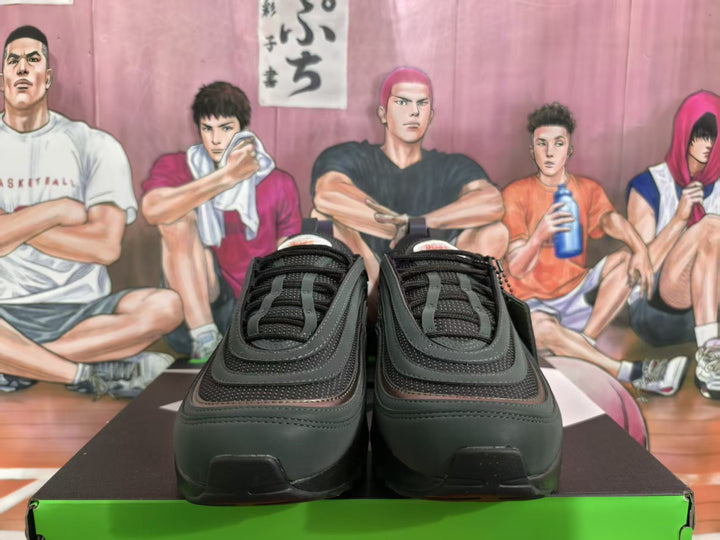 Pair of black and dark green sneakers shown from the front on a green shoebox, with layered uppers and green accents.