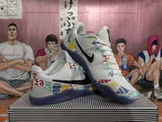 Angled view of white basketball shoes with black Swooshes, colorful geometric graphics and dark blue outsoles.