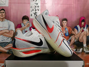 Side view of white Nike basketball sneakers with black and orange-red Swooshes, perforated panels, and a speckled midsole.