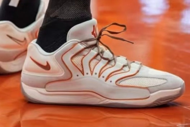 White and orange Nike low-top sneaker with mesh panels, layered side detailing, brown laces, and a rubber outsole on an orange surface.
