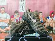 Pair of brown knit Nike sneakers displayed on a shoebox, showing black patterned detailing, contrasting Swooshes, and textured black outsoles.